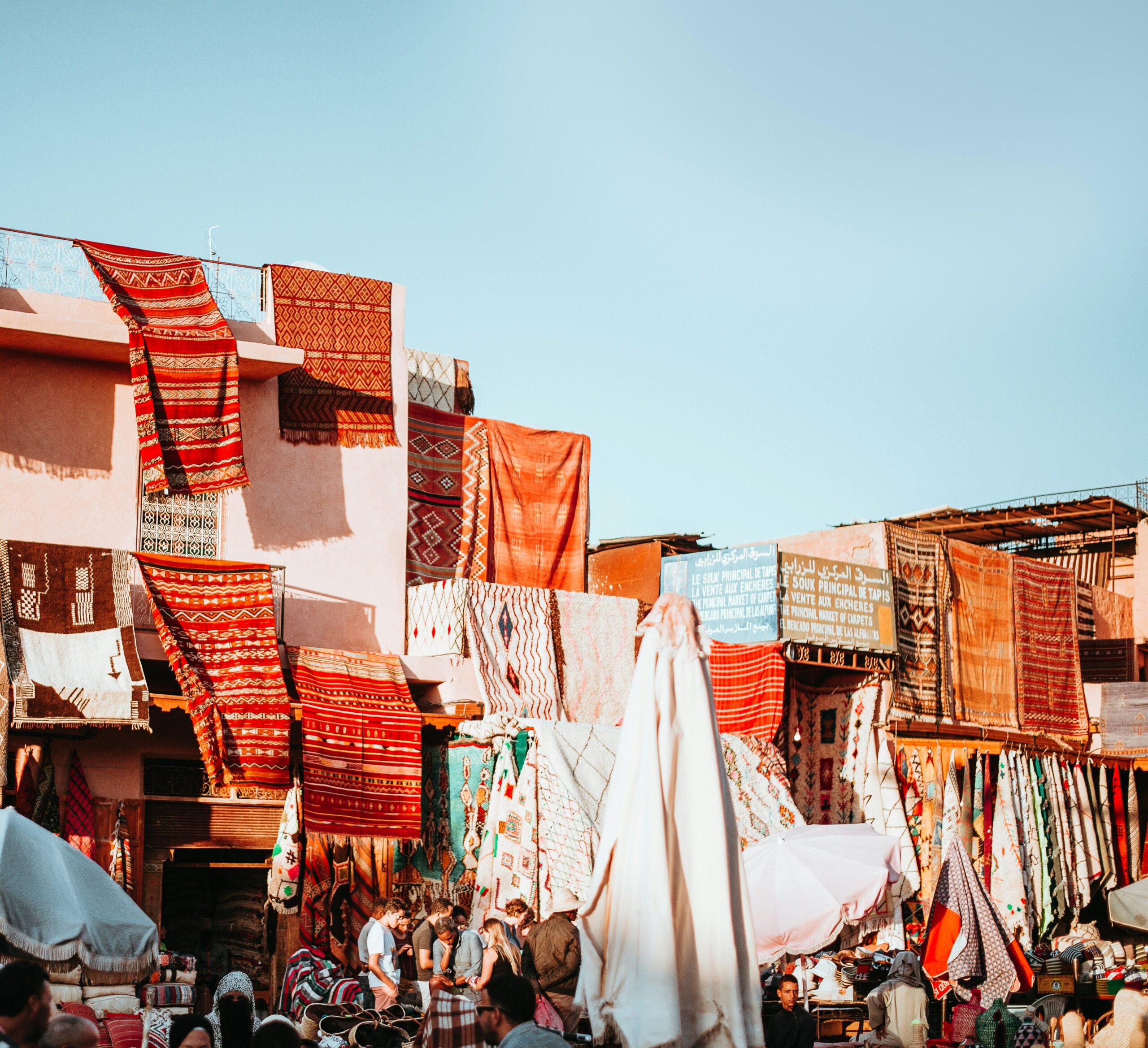 A lively outdoor market with traditional handwoven rugs hanging from the walls of pastel-colored buildings