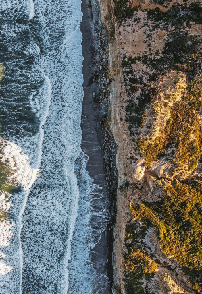  Aerial view of a rugged coastline where the ocean waves crash against a narrow beach, bordered by steep cliffs 